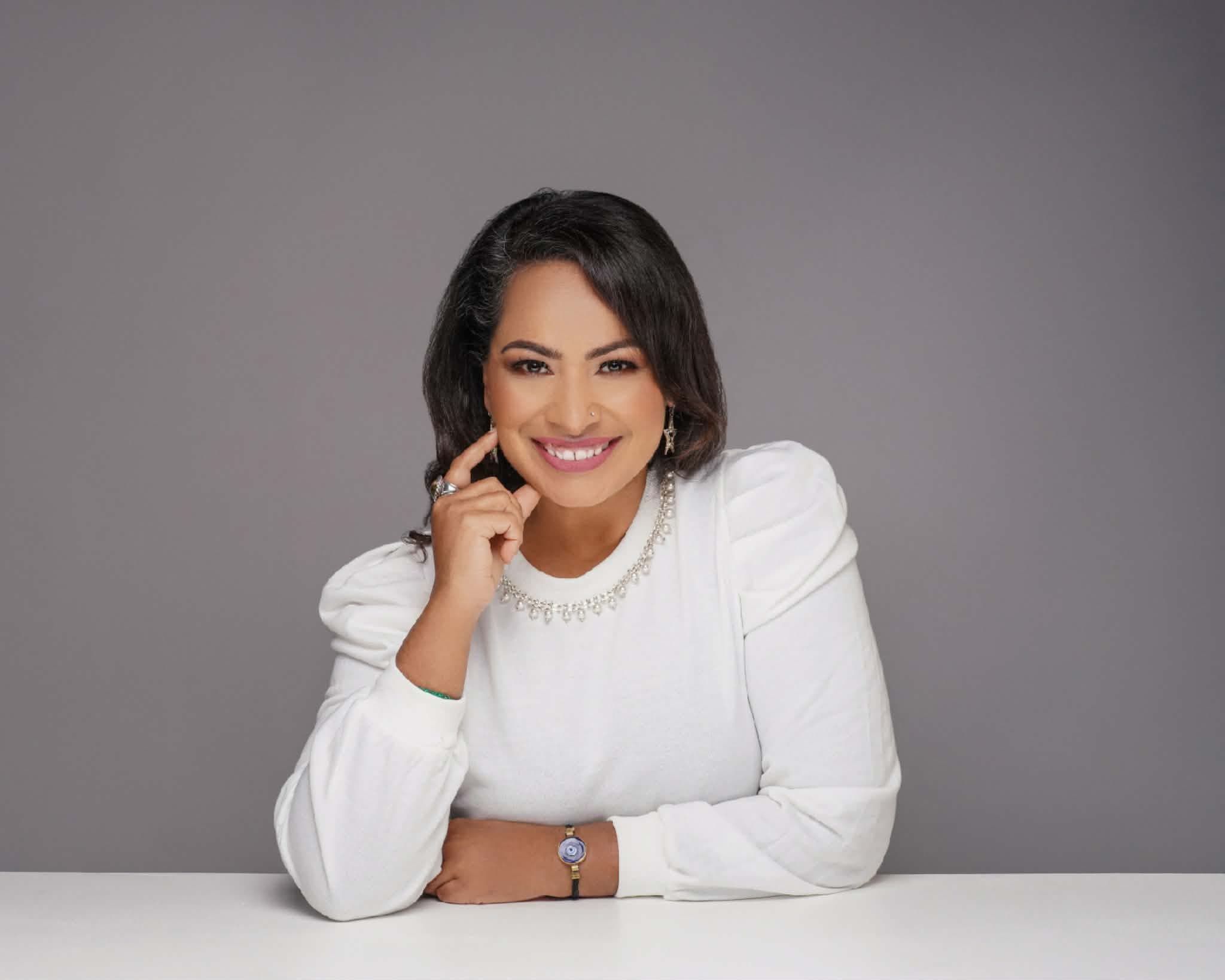 Portrait of Dina Sabnani smiling in a white sweater with a jeweled collar, seated against a soft gray backdrop